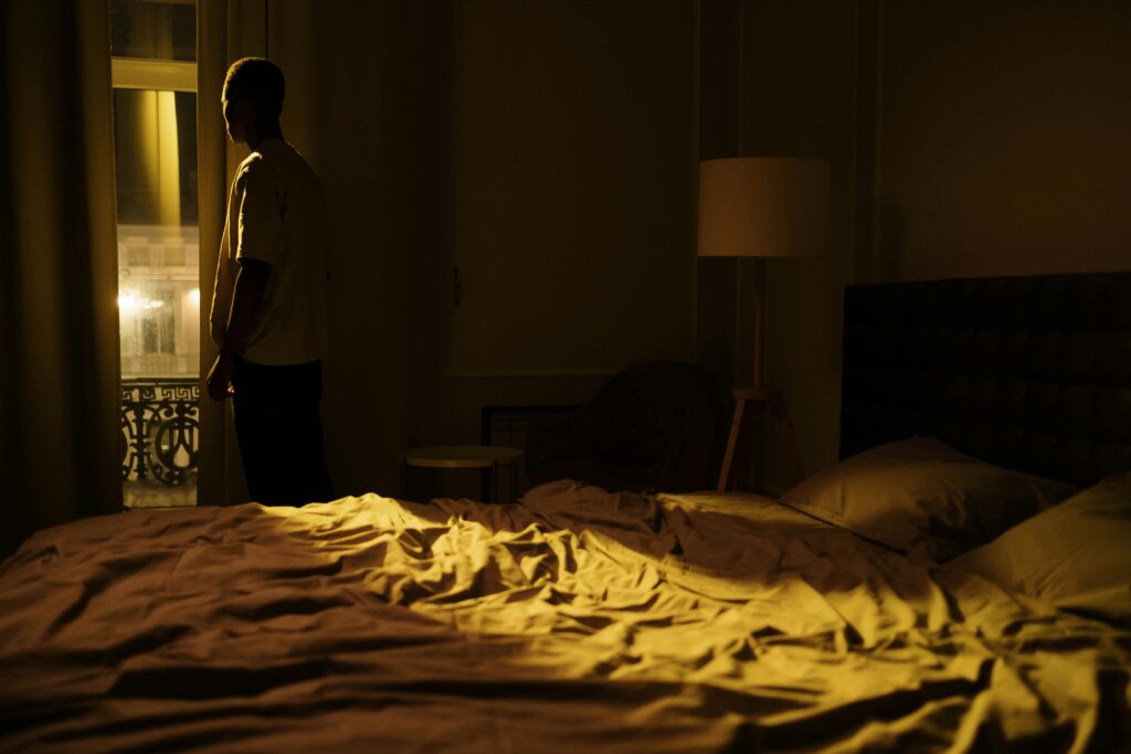 Man in dark bedroom gazing out window, highlighting themes of insomnia and solitude.